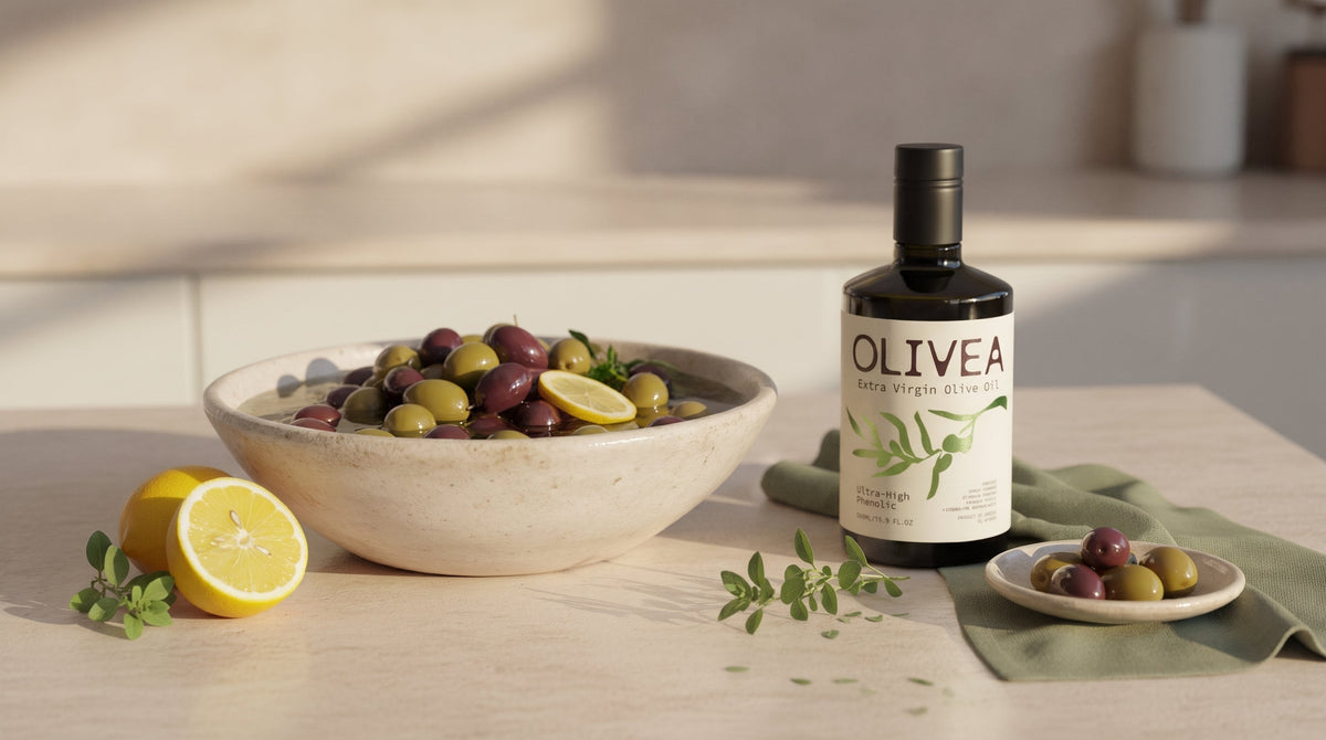 A ceramic bowl of green and purple table olives in brine on a limestone counter beside a bottle of Olivea extra virgin olive oil, with lemon and fresh oregano