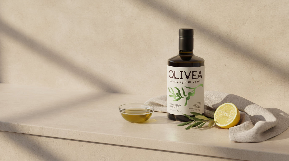 A bottle of Olivea extra virgin olive oil on a sunlit limestone vanity with a glass bowl of oil, a folded linen towel, an olive branch and half a lemon